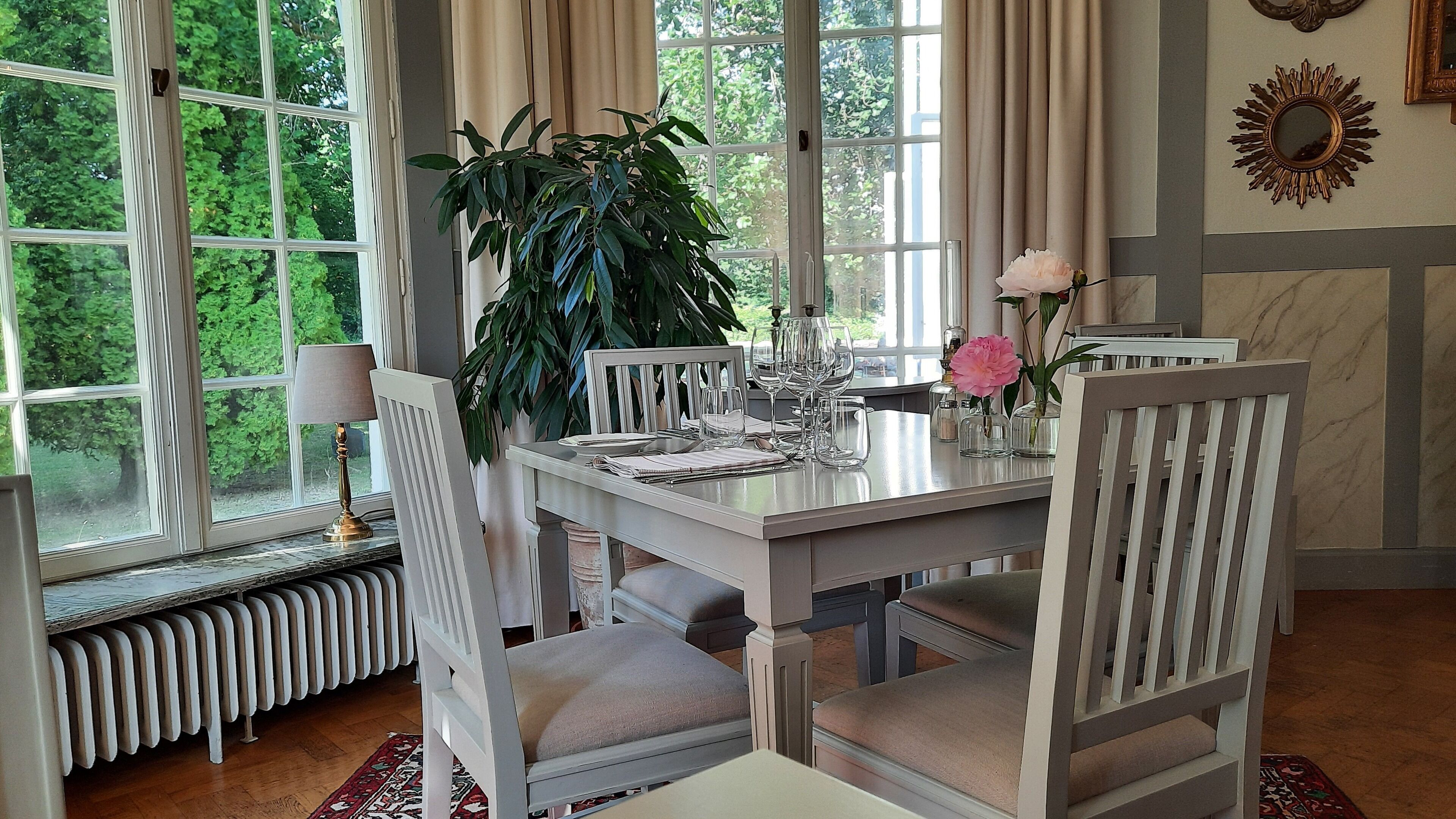 Un coin repas avec une table dressée pour quatre personnes, entourée de chaises blanches dotées d’assises rembourrées. Un pot de fleurs est posé sur la table, ainsi que des objets décoratifs, notamment un vase avec une fleur rose. La pièce est dotée de grandes fenêtres avec des rideaux, et un radiateur est visible sous la table.