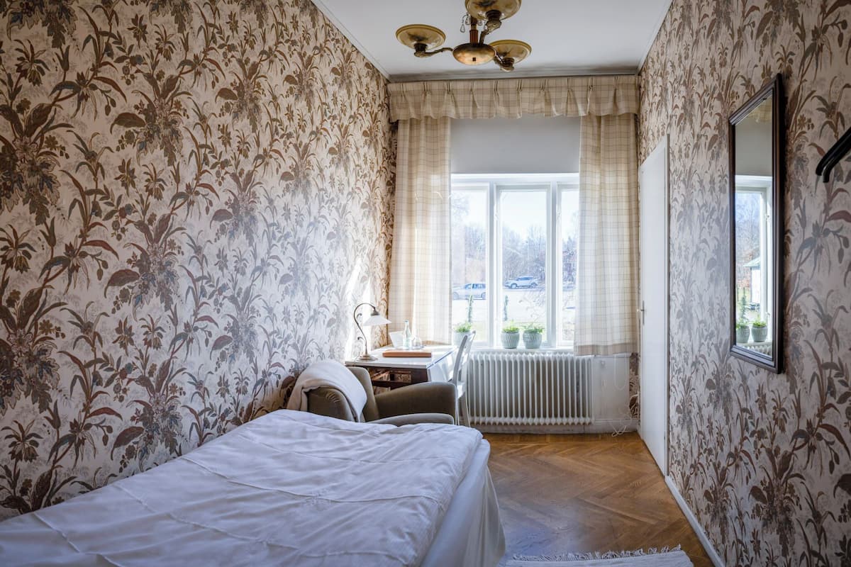 A bedroom with floral wallpaper, a bed, a desk with a chair, a radiator, and a window with curtains.