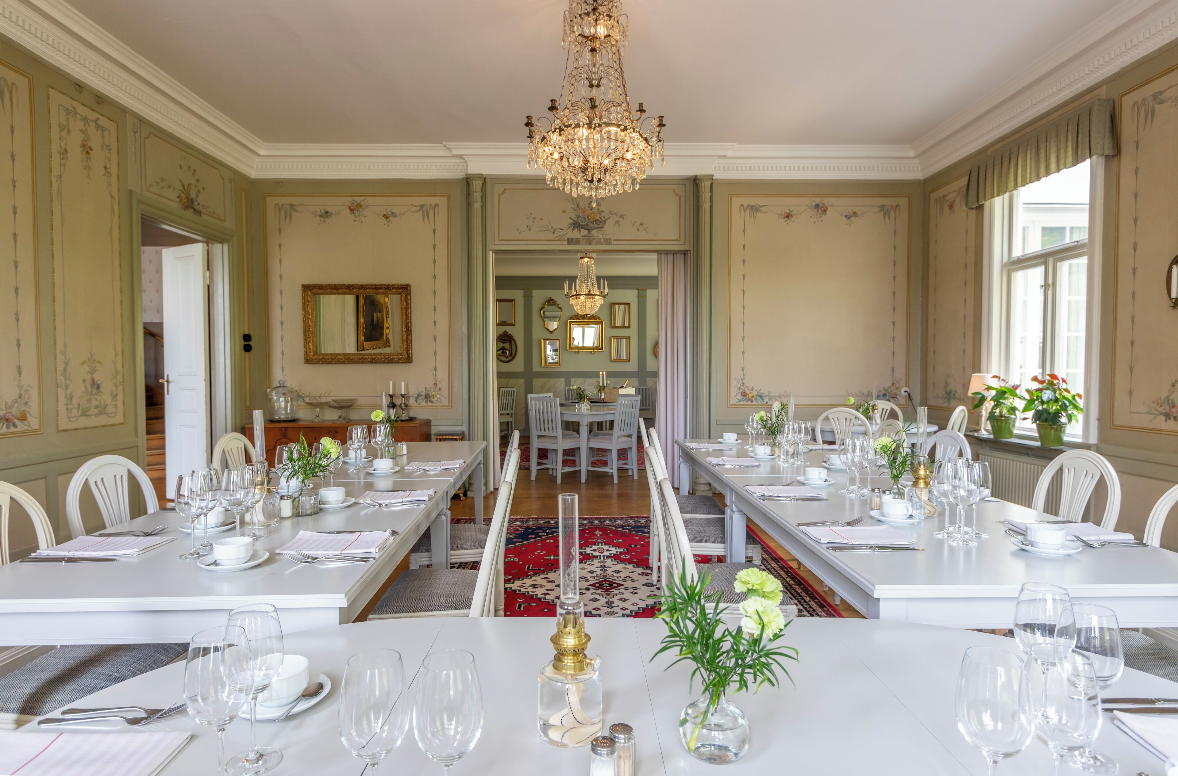 Une salle à manger élégante, avec de longues tables dressées pour un repas, un lustre et une vue sur un espace de restauration.