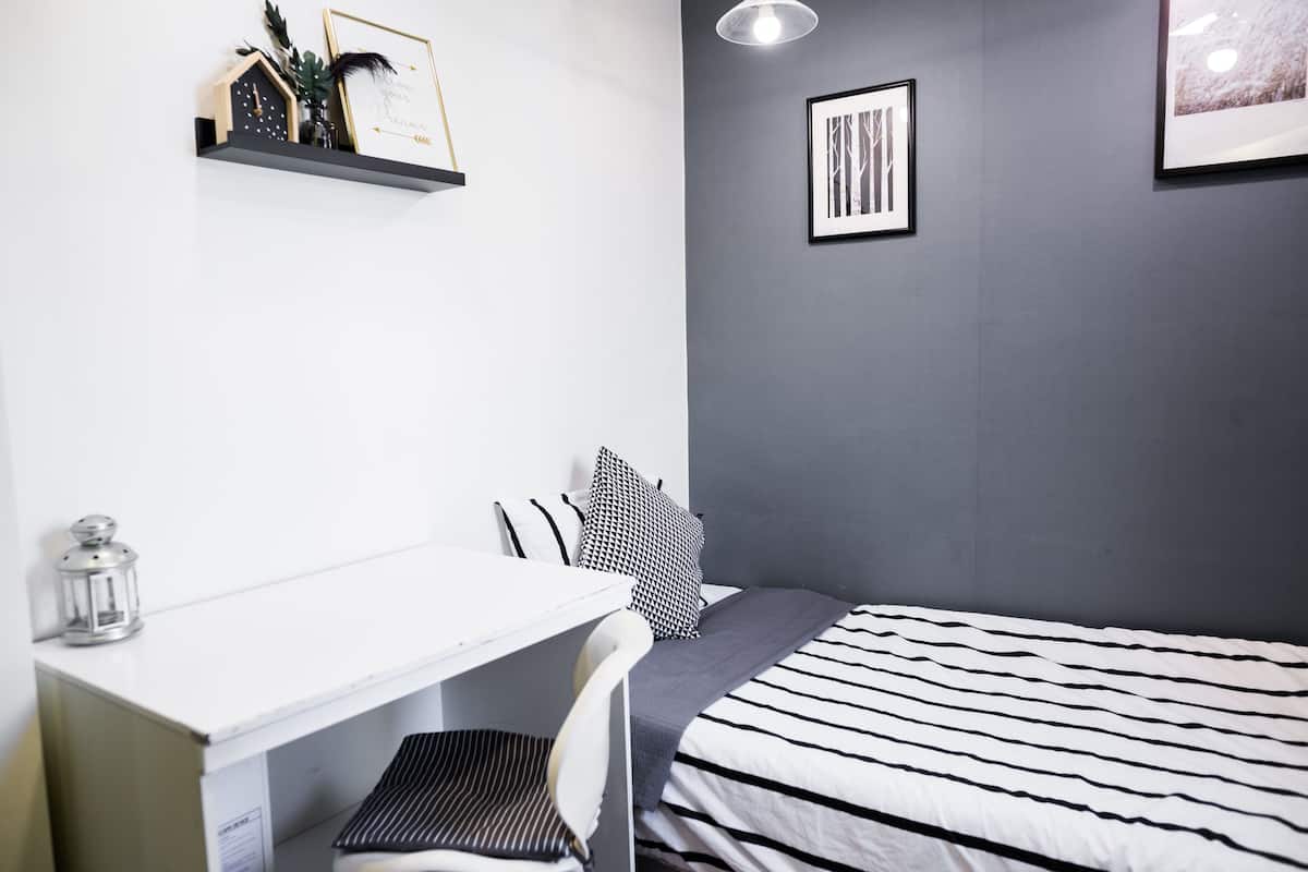 A bedroom with a bed, a desk, and a chair. There is a shelf with decorative items and a framed picture on the wall.