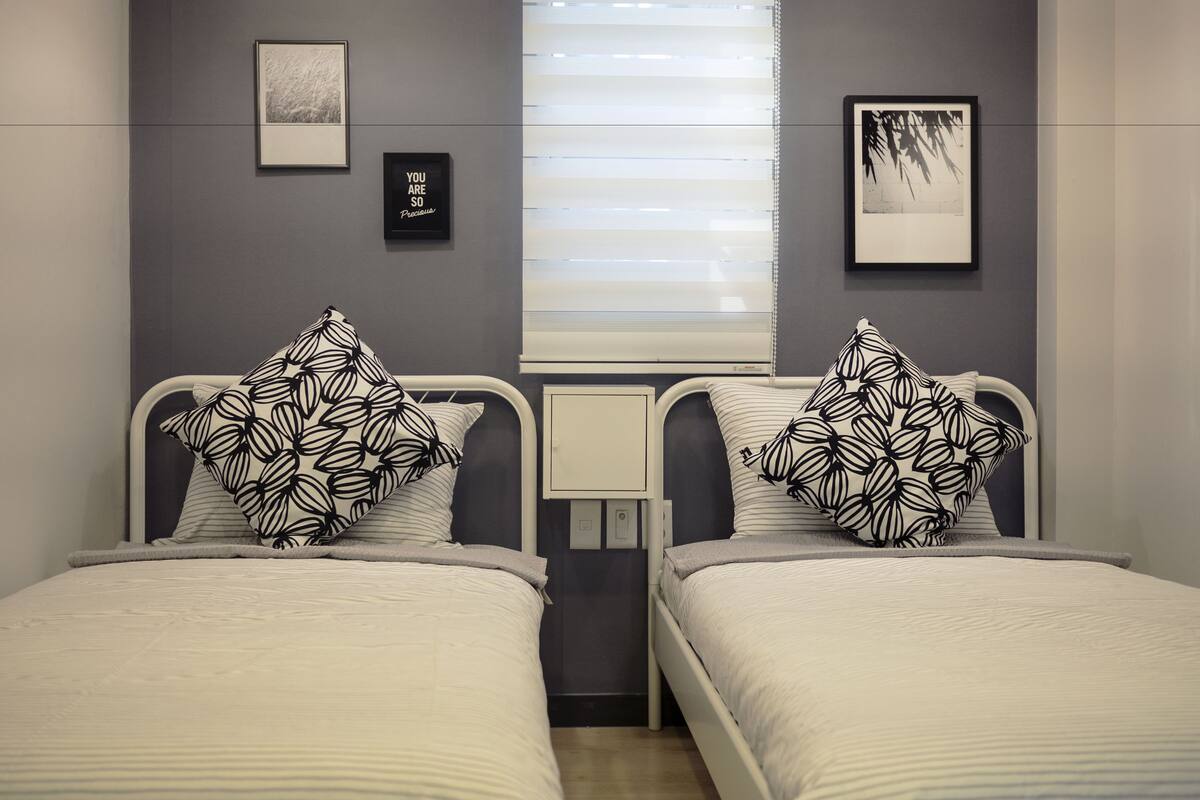 A twin room with two beds, each with a patterned pillow, a bedside table, a framed picture on the wall, and a window with blinds.