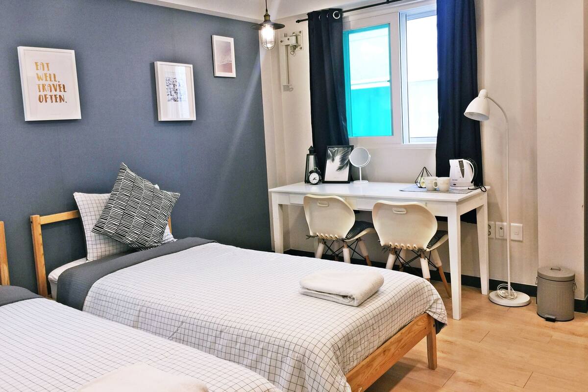 A modern bedroom with a bed, a desk, and a chair. There is a window with curtains, a lamp, and a trash bin.
