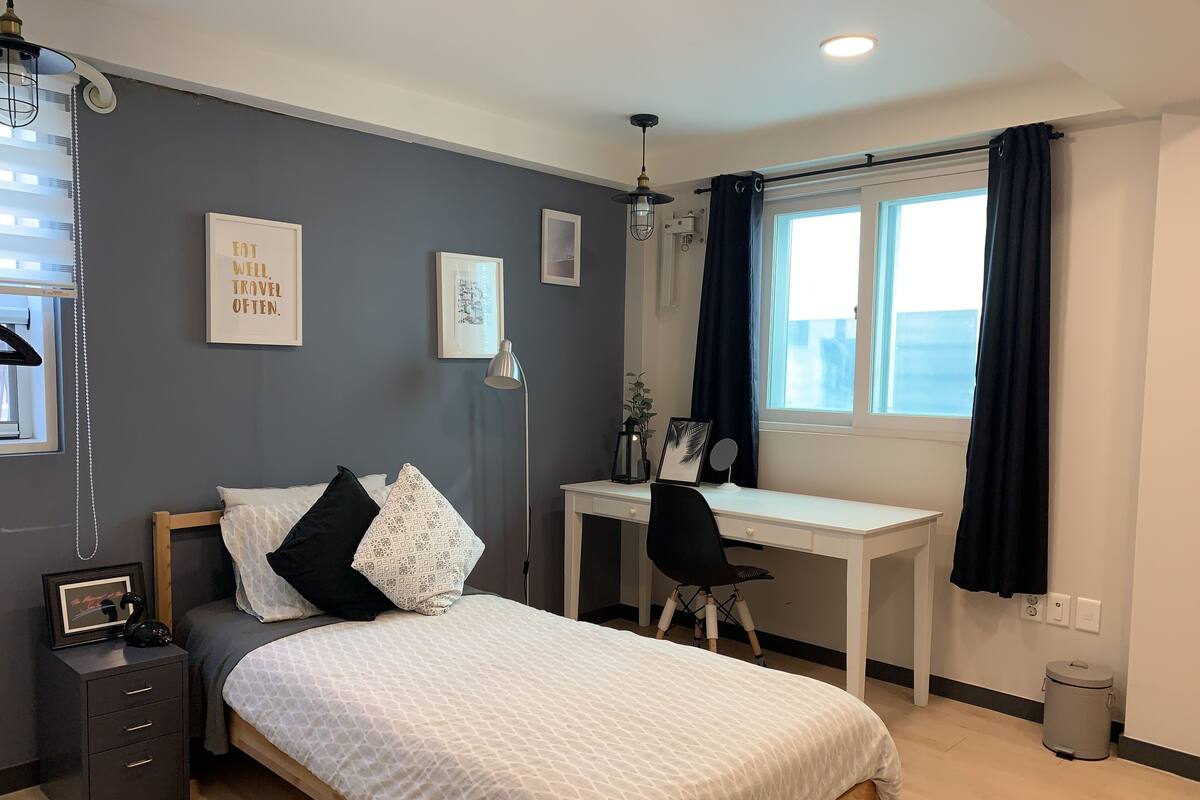 A bedroom with a bed, bedside table, desk, chair, and a window with curtains.