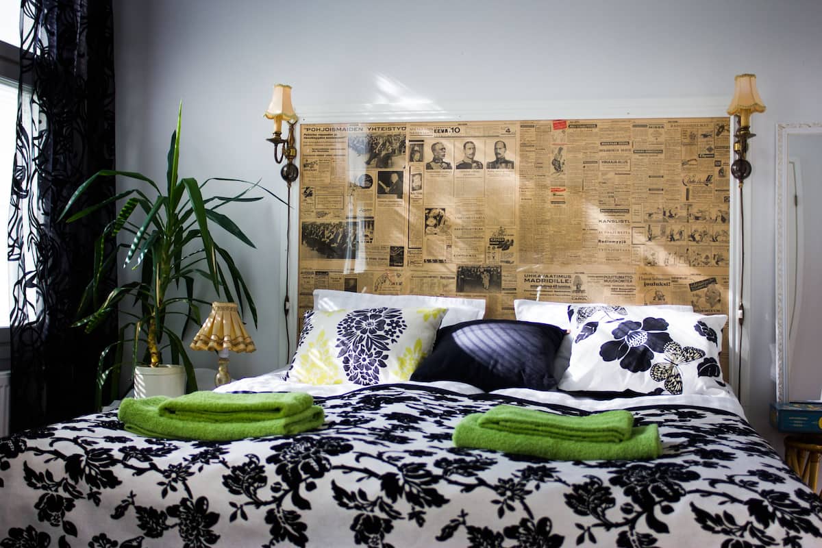 Ein Bett mit floralem Bettzeug, ein Kopfteil mit Zeitungsausschnitten, eine Topfpflanze und eine Lampe.