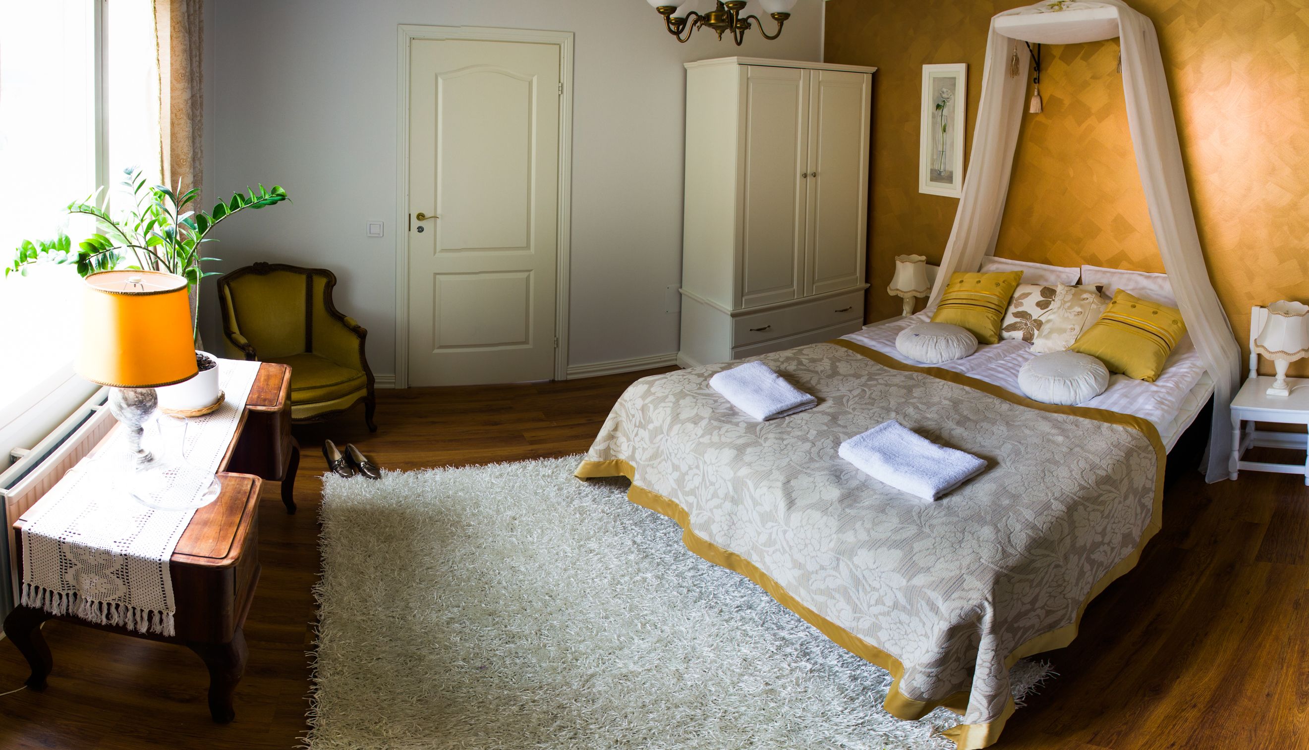 Un dormitorio con cama, una silla, un escritorio con lámpara, un armario y una ventana con cortinas.