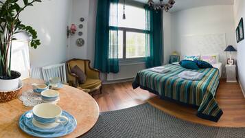 A well-lit room with a bed, a chair, a table with cups and saucers, and a potted plant.
