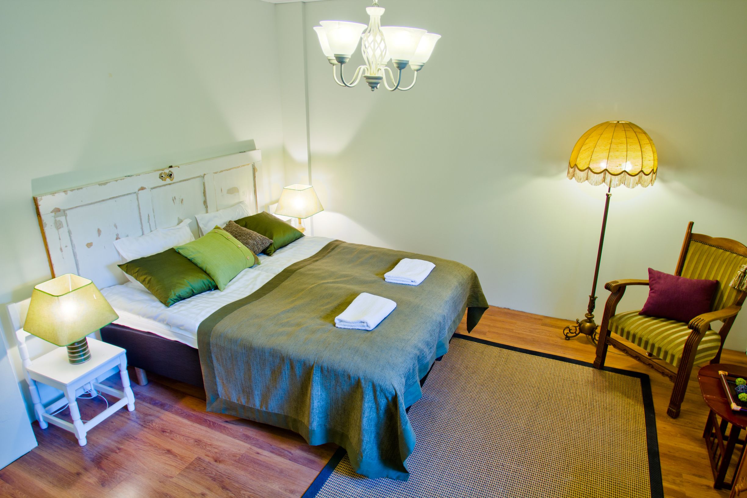 Una cama bien hecha con una colcha verde, dos almohadas y dos toallas dobladas. Hay una mesita de noche con una lámpara, una silla con un cojín y una lámpara de pie con pantalla amarilla.