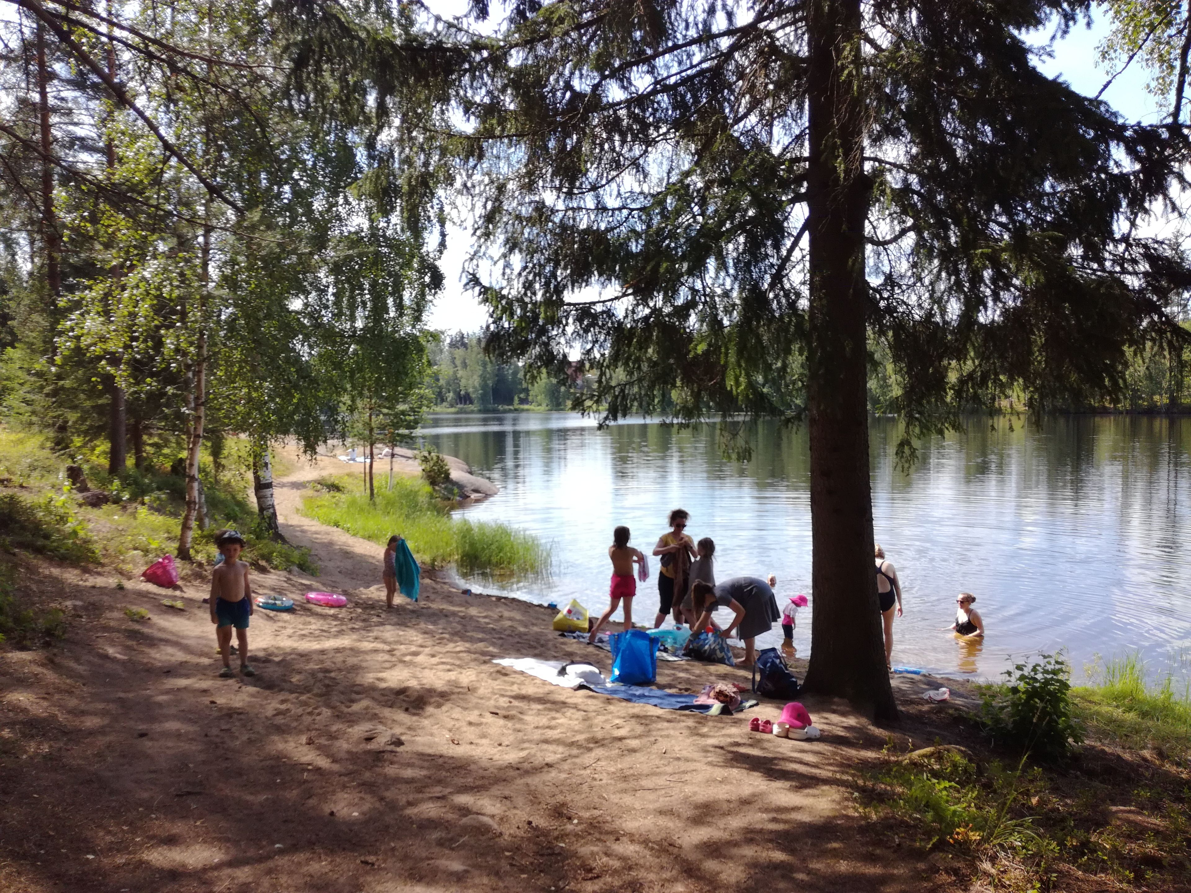 A lakeside campsite with trees, a sandy area, and people engaging in various activities.