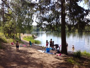 A lakeside campsite with trees, a sandy area, and people engaging in various activities.