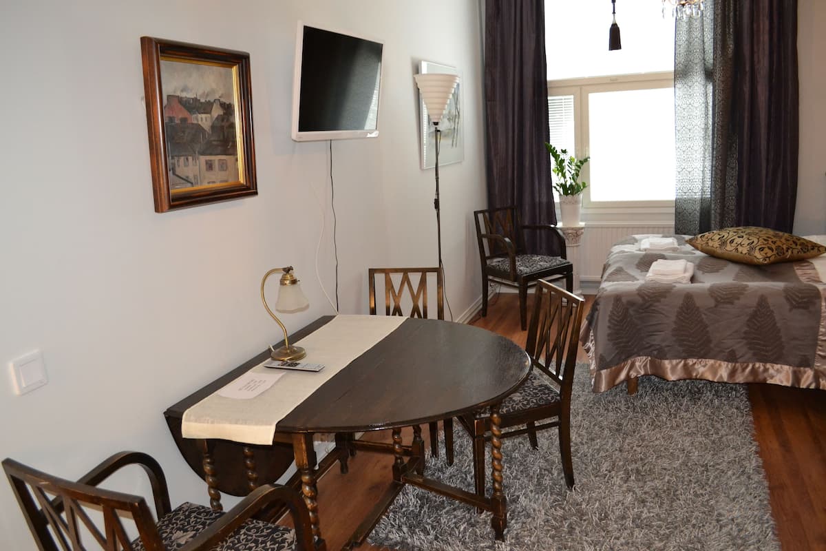 Ein Zimmer mit Esstisch, Stühlen, einem Bett mit gemustertem Überwurf, einem Fernseher und einer Lampe.