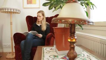 A person sitting on a red velvet chair reading a book in a room with a floor lamp, a potted plant, and a patterned rug.