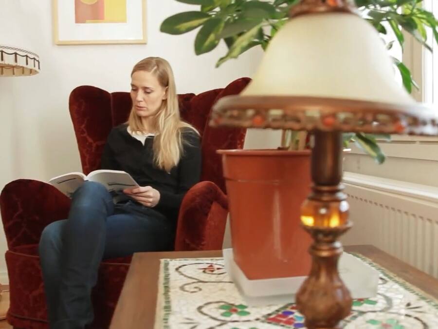 A person sitting on a red velvet chair reading a book in a room with a floor lamp, a potted plant, and a patterned rug.