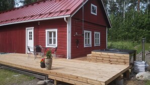 Punainen puutalo, jossa on terassi, pieni pöytä tuoleineen ja ruukussa oleva kasvi.