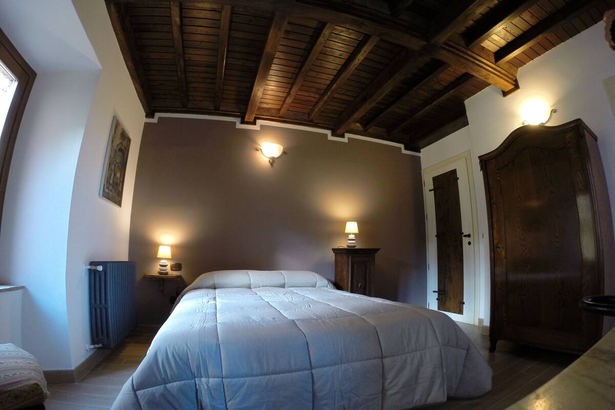 Een slaapkamer met een groot bed, houten balken in het plafond, twee bedlampen, een radiator en een kledingkast.