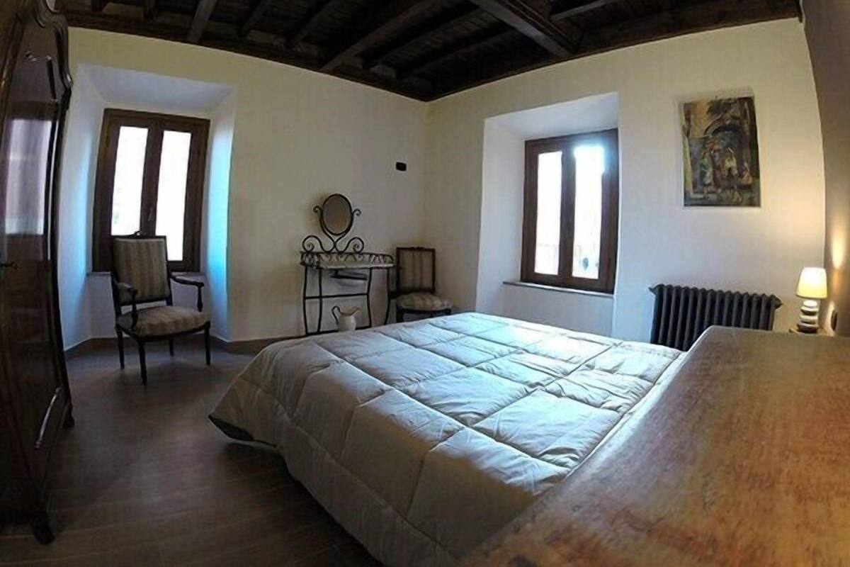 Een slaapkamer met een bed, een stoel, een klein tafeltje met een spiegel, en twee ramen.