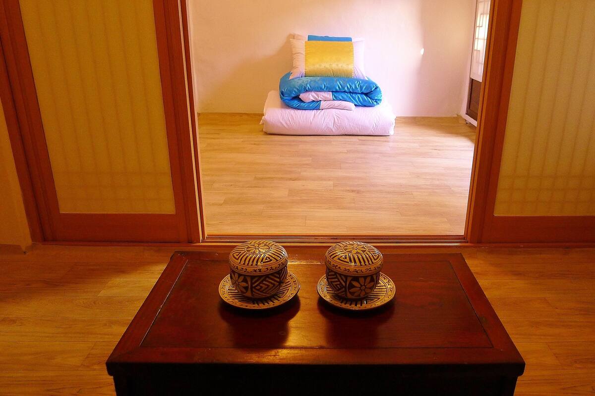 A room with a wooden floor, a small table with two decorative items, and a view of a bed with pillows and a blanket.