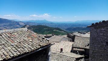 Vista di una città storica con edifici in pietra e tetti in terracotta, affacciata su una valle e montagne lontane.