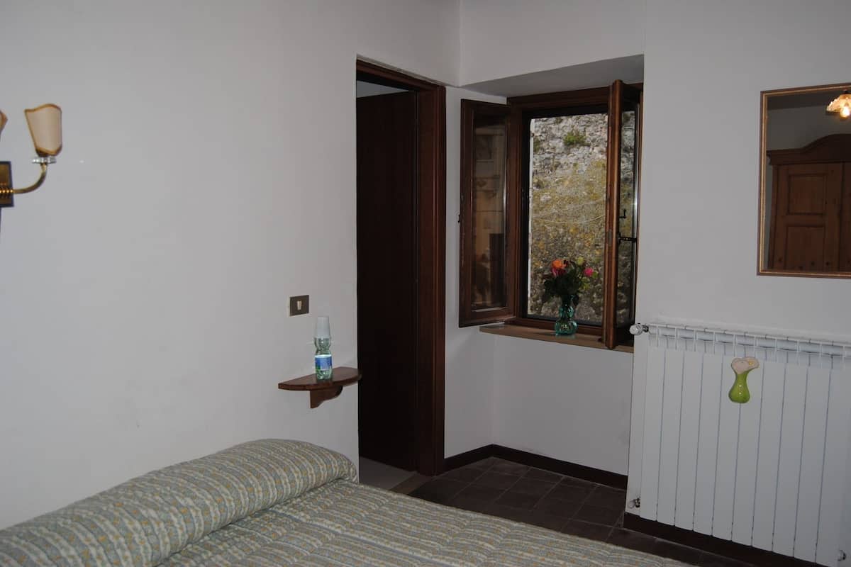 A small, cozy room with a bed, a window with a view of a stone wall, a radiator, and a shelf with a bottle.
