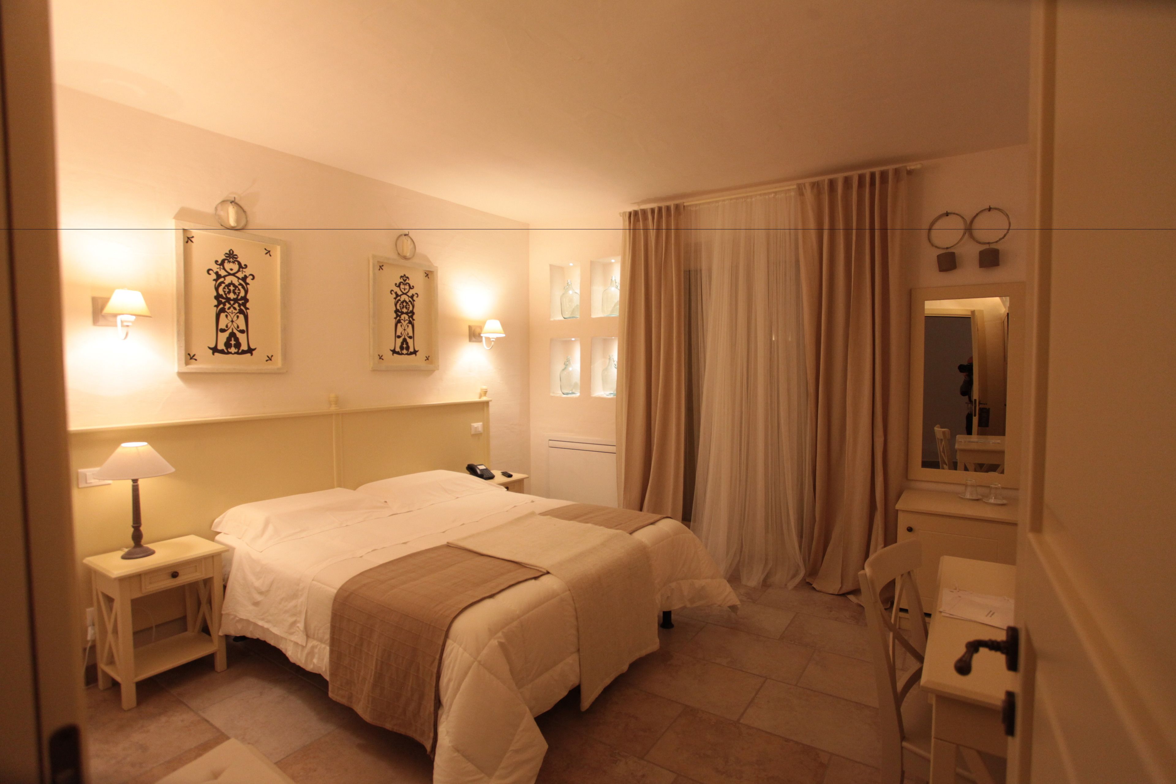 A hotel room with a large bed, bedside lamps, a nightstand, a desk with a chair, a mirror, and a window with curtains.