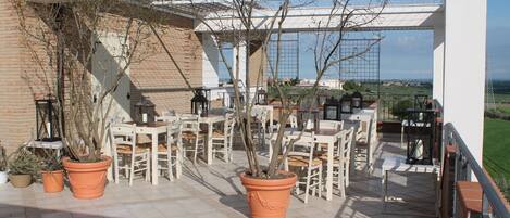 An outdoor patio with potted plants, white chairs, and tables, overlooking a scenic landscape.