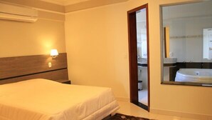 A hotel room with a bed, a nightstand, a lamp, a mirror, and a view of a bathroom with a bathtub. - Terraço Hotel (Tres Pontas)