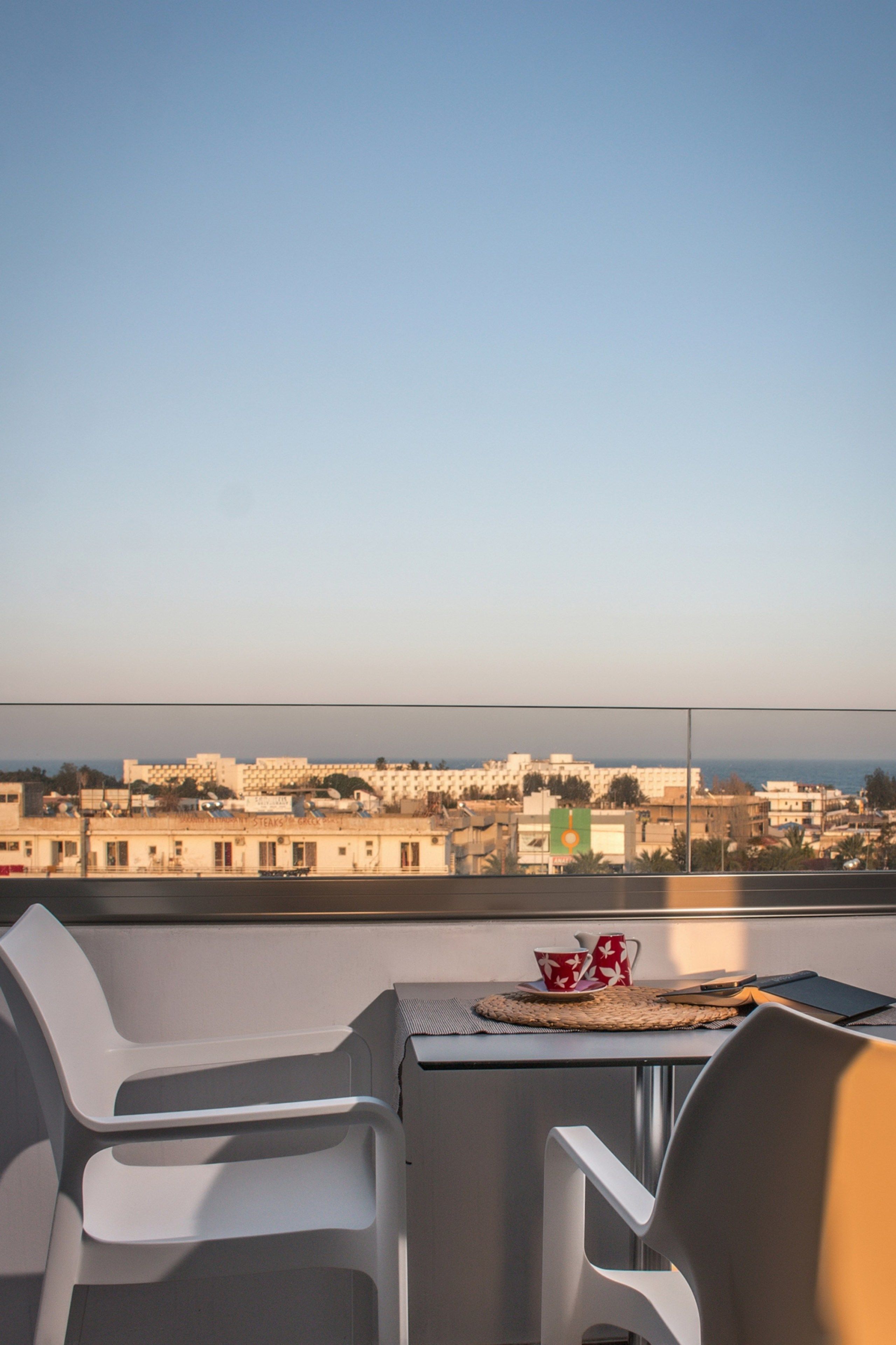 A balcony with two white chairs, a table, and a view of a cityscape with buildings and a body of water.