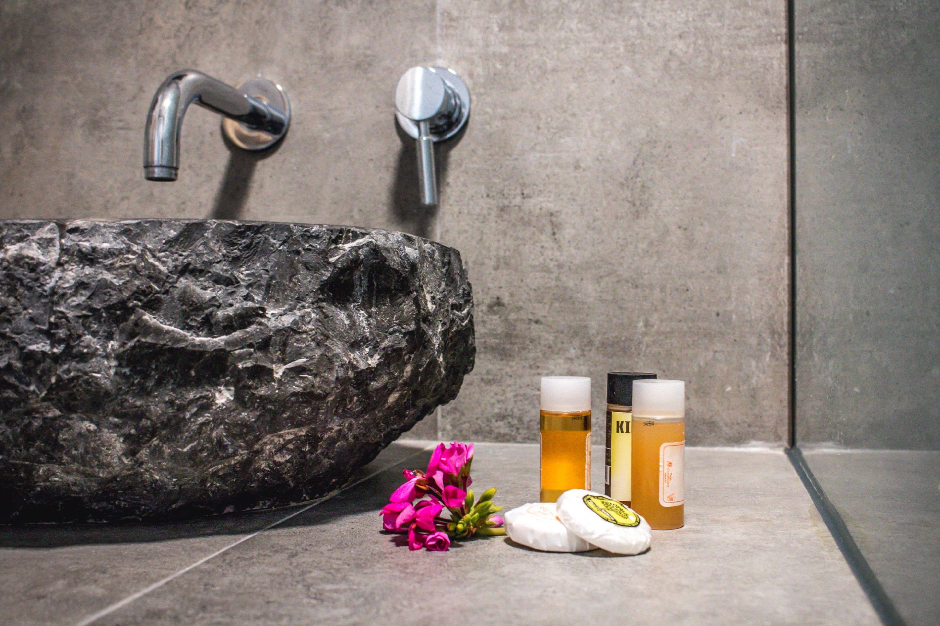 A bathroom with a large, textured stone sink, two chrome faucets, and a bottle of lotion and a bottle of shampoo.