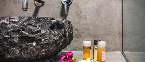 A bathroom with a large, textured stone sink, two chrome faucets, and a bottle of lotion and a bottle of shampoo.