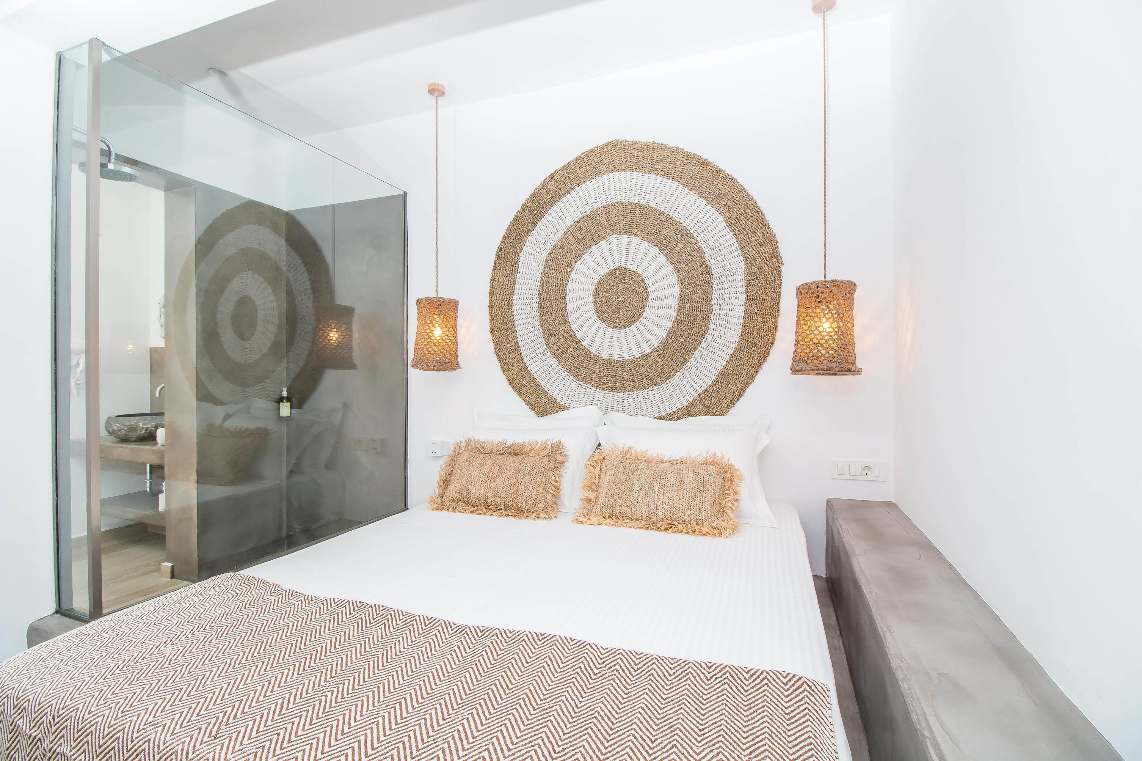 A modern bedroom with a glass shower enclosure, a large circular woven wall art piece, and two hanging pendant lights.