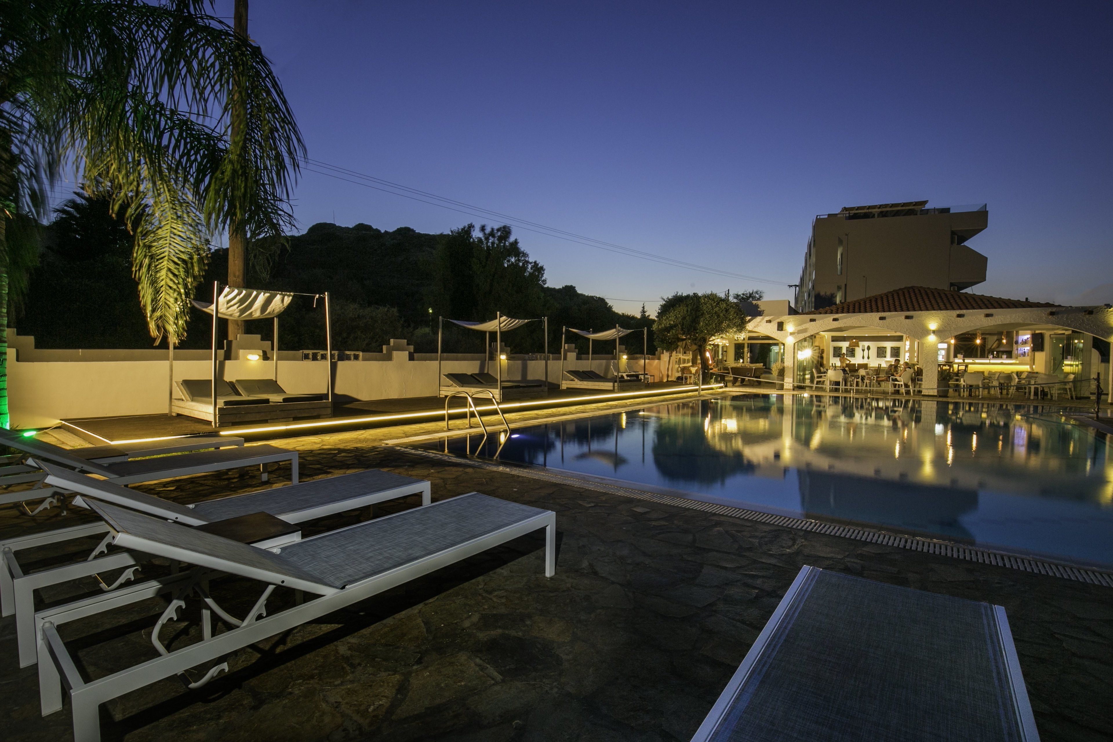 Un espace piscine avec des chaises longues et des parasols, un bâtiment doté d’une architecture originale et un ciel nocturne dégagé.