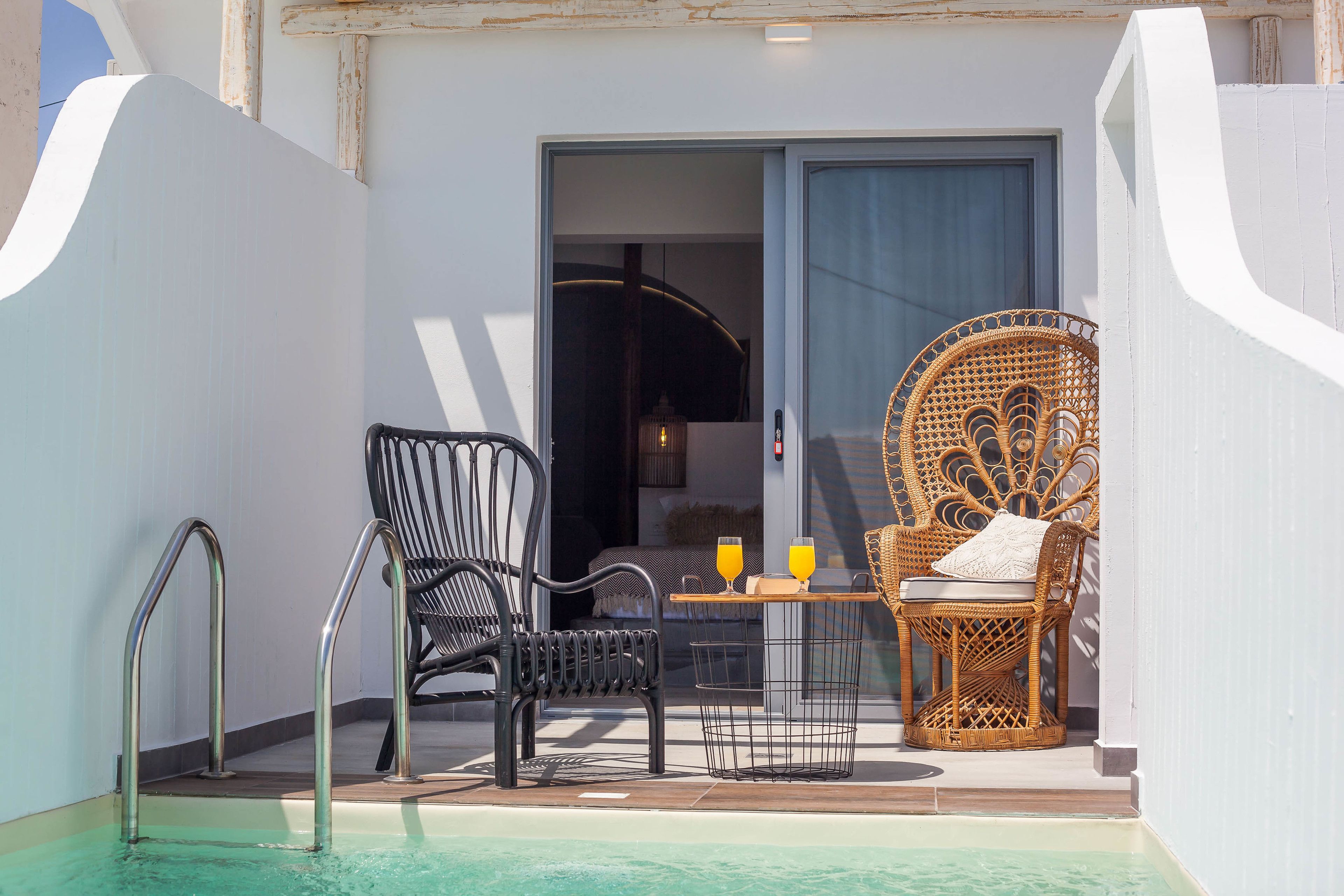 A balcony with a pool, two wicker chairs, a small table with drinks, and a door leading to a room.