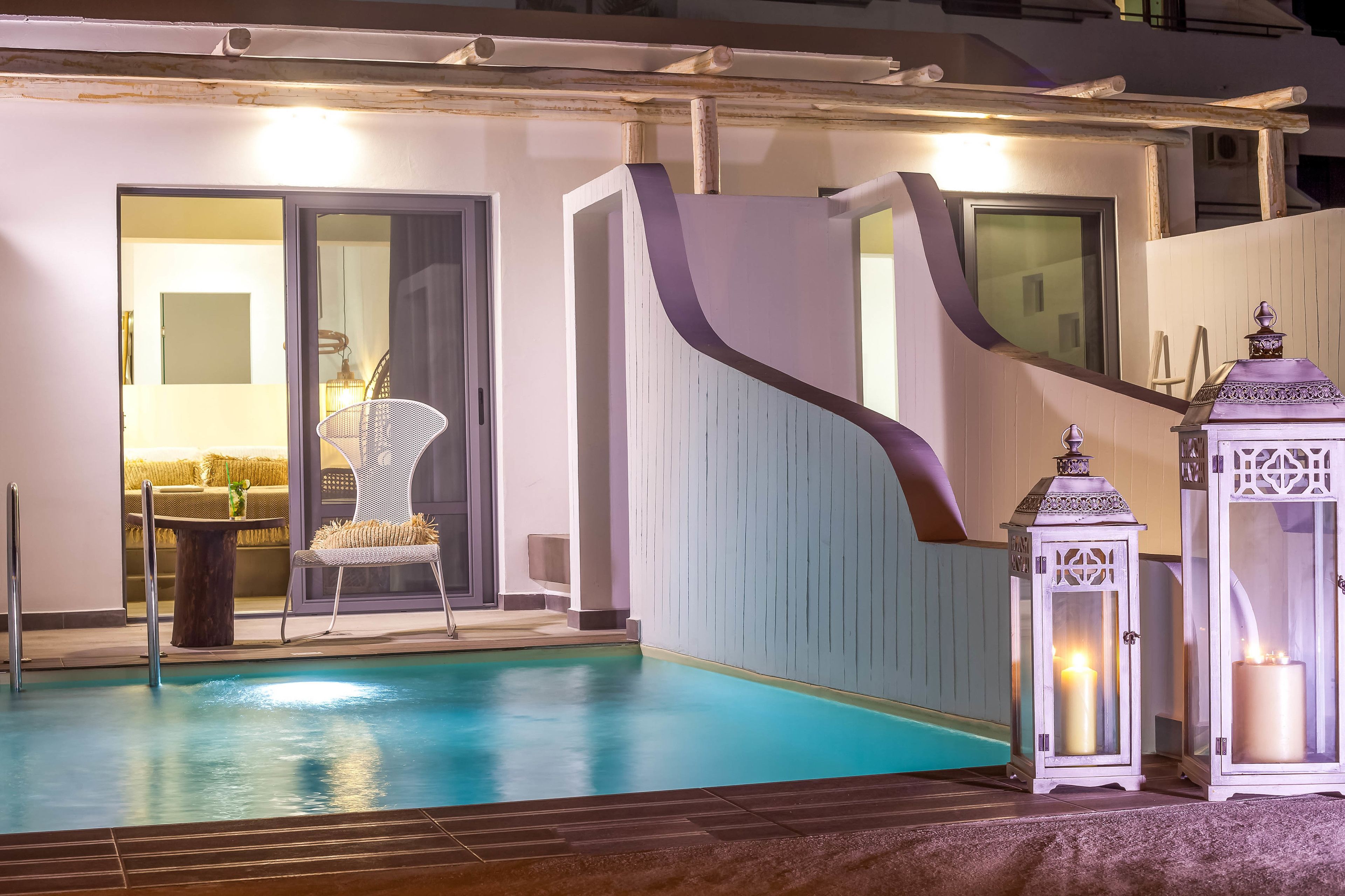 Una piscina con un muro curvo, una silla y dos faroles iluminados.