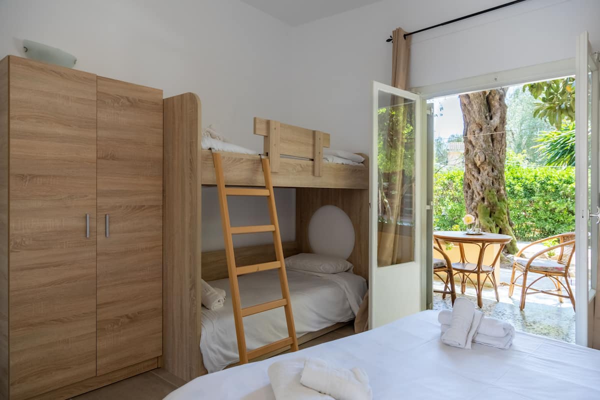 A room with a bunk bed, a wooden wardrobe, a ladder, a bed with towels, and a view of an outdoor area with a table and chairs.