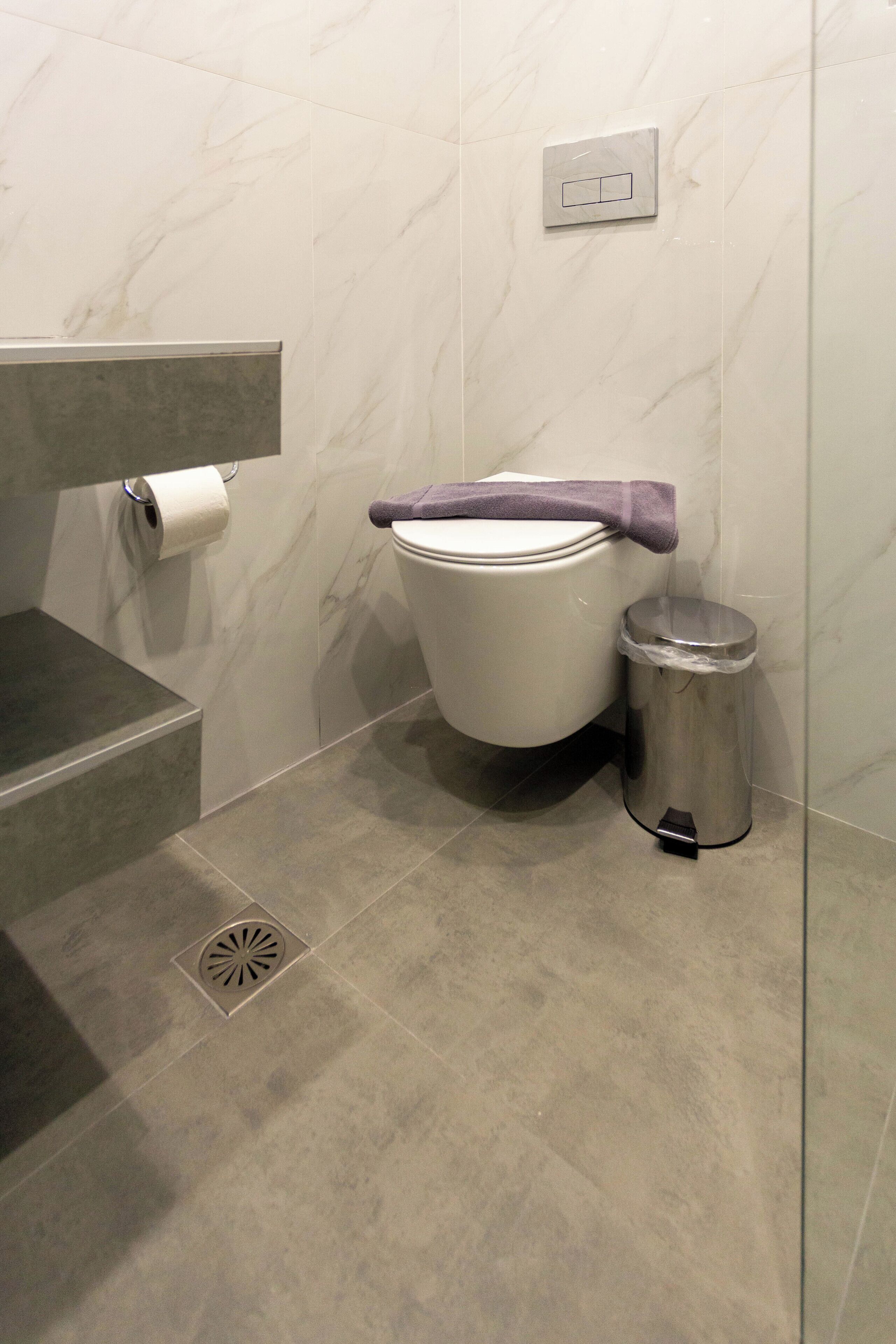 Un baño moderno con inodoro, un cubo de basura, pared de mármol y desagüe de ducha.