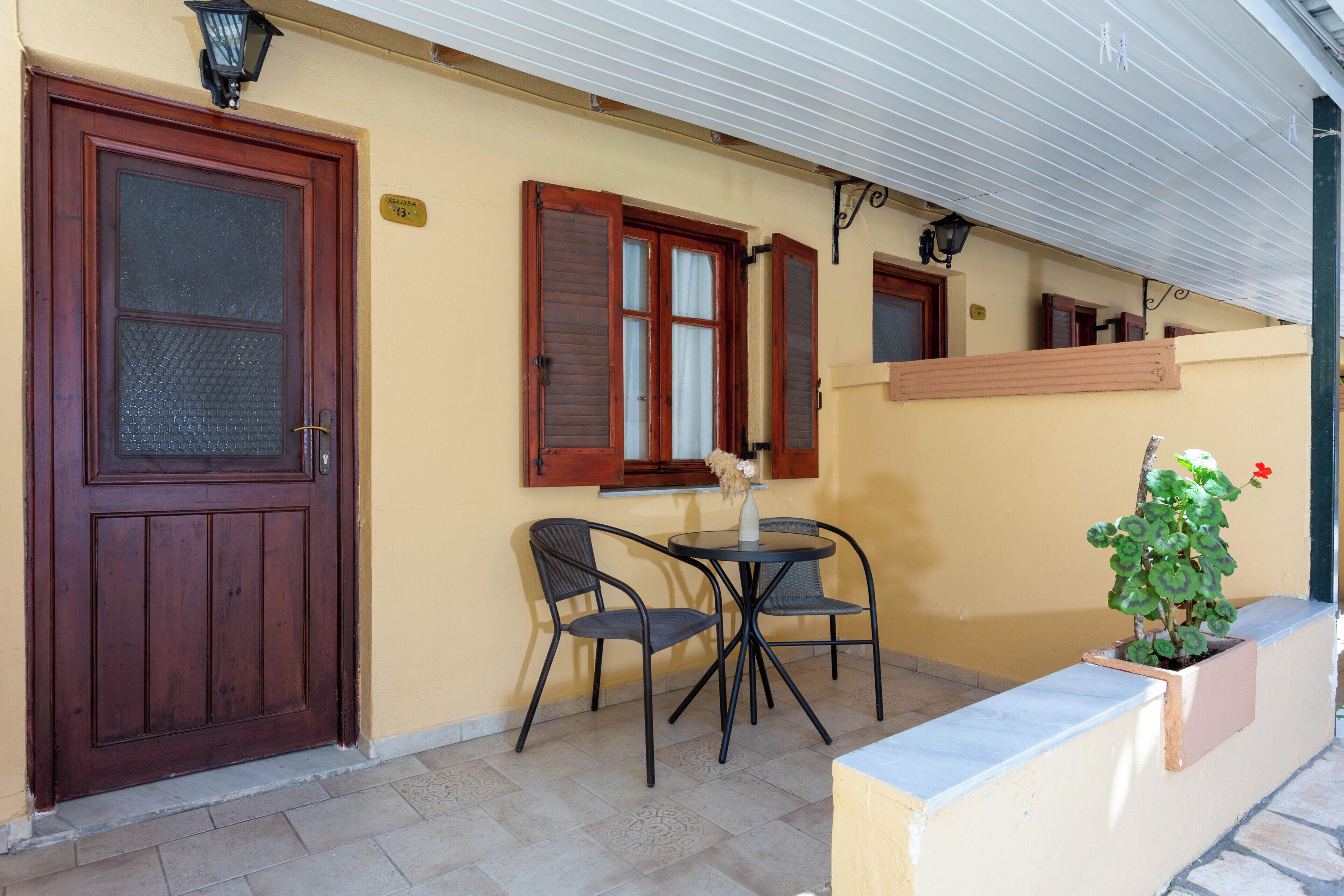 A building with a wooden door, a window with shutters, a small table with chairs, and a potted plant.