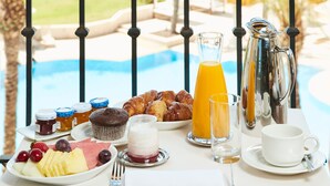 Una mesa de desayuno con cruasanes, fruta, tarros de miel, un vaso de jugo de naranja, una jarrón, una taza y un platillo junto a una piscina.