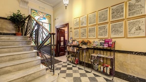 Un hall d’hôtel avec un sol à carreaux, un escalier doté d’une rampe en métal, un mur orné de cartes encadrées et un comptoir de réception agrémenté de livres et d’une plante en pot.