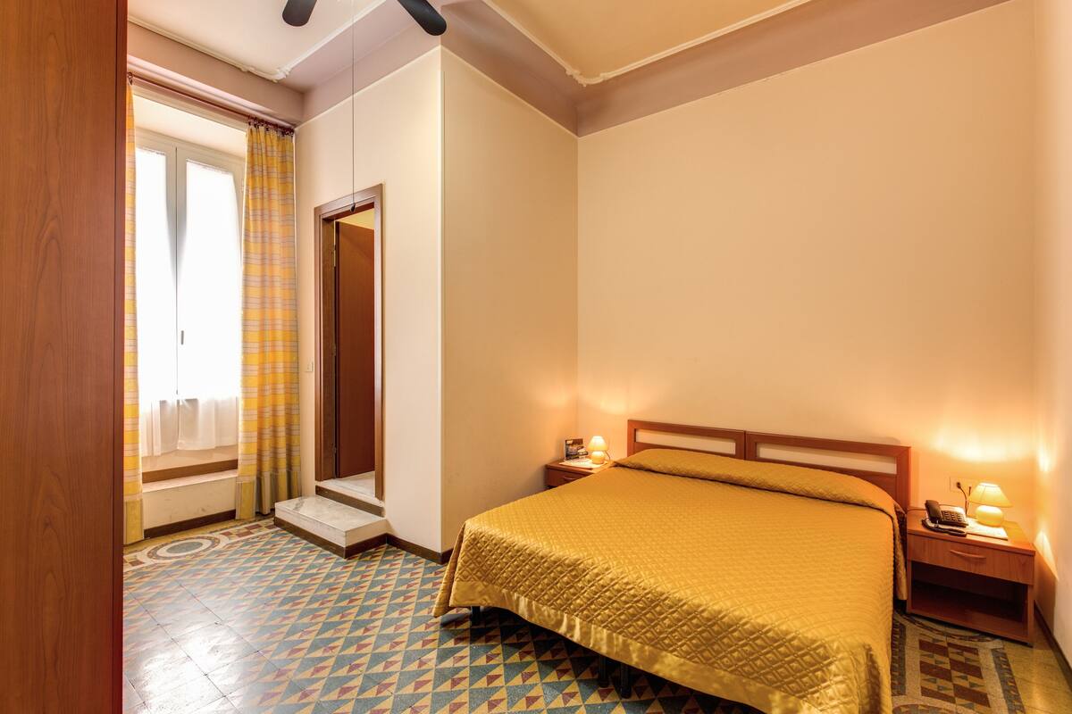 Een hotelkamer met een bed, een nachtkastje met een lamp, een telefoon en een raam met gordijnen.