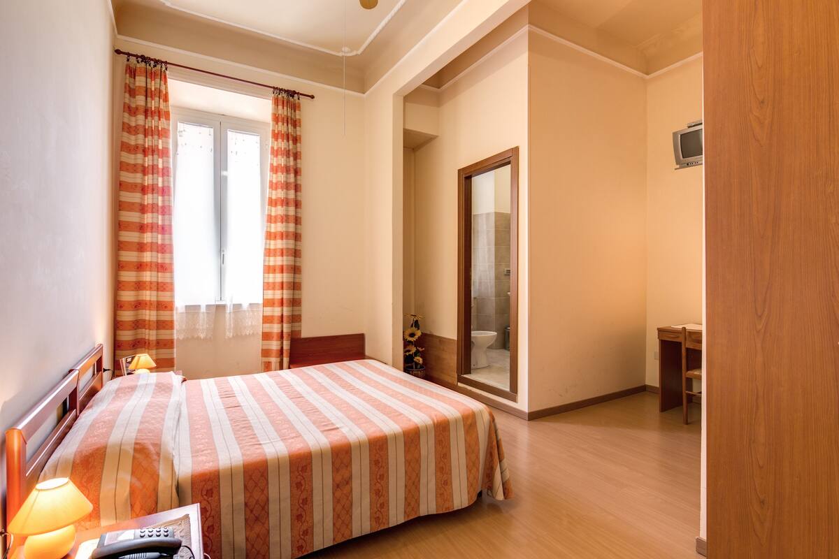 Een hotelkamer met een bed, nachtkastje met lamp, klein bureau en douche.