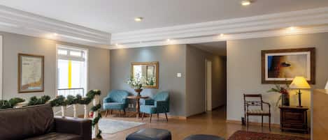 Amplia sala de estar con un sofá de cuero marrón, dos otomanas azules, una mesa de centro y suelo de madera. Hay dos sillones turquesa, una mesita de noche con una lámpara y un cuadro enmarcado en la pared.