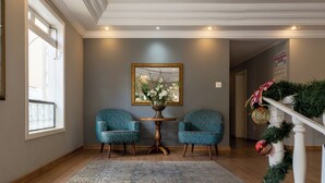 Una habitación con dos sillones estampados, una pequeña mesa redonda con un jarrón, un ventanal grande y una escalera decorada con plantas.