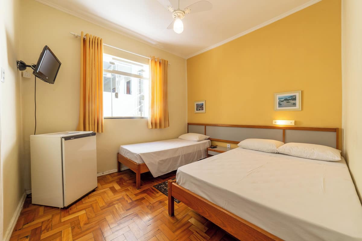 A hotel room with two single beds, a double bed, a small refrigerator, a ceiling fan, and a television on the wall.