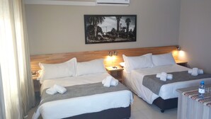 A hotel room with two beds, a nightstand, a table with a bottle and glasses, and a painting on the wall. - Prost Hotel (Swakopmund)