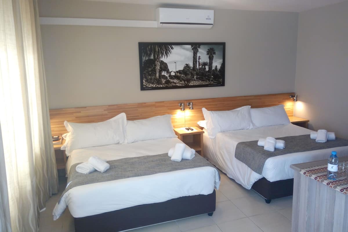 A hotel room with two beds, a nightstand, a table with a bottle and glasses, and a painting on the wall.