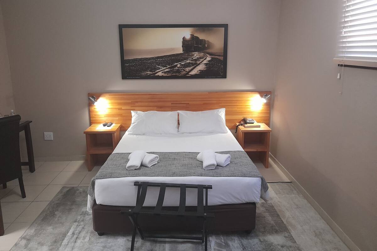 A hotel room with a bed, two bedside tables, a headboard, a nightstand, a chair, and a painting on the wall.