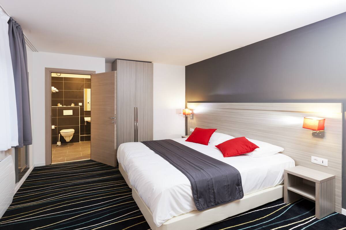 Une chambre d’hôtel moderne avec un grand lit, une table de chevet, une petite table d’appoint, une lampe fixée au mur et une salle de bain visible à travers une porte ouverte.