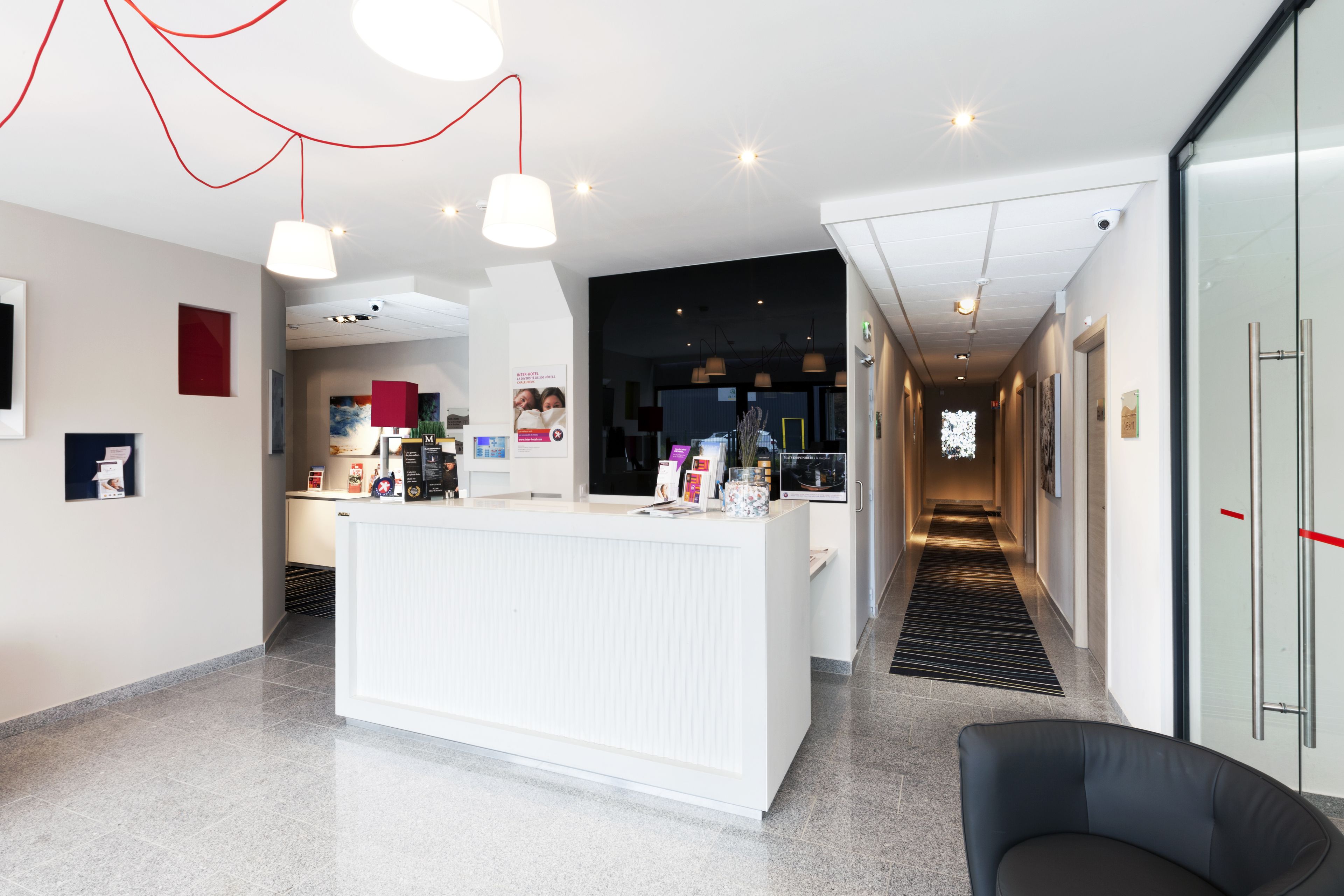 Een moderne hotellobby met een receptiebalie, een zwarte leren fauteuil en een glazen deur aan de rechterkant.