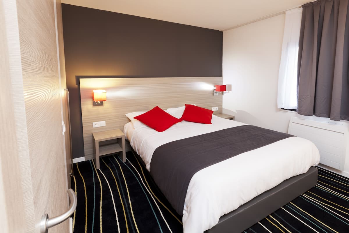 Une chambre d’hôtel avec un lit, une table de chevet, une lampe fixée au mur, une fenêtre avec des rideaux et un sol à rayures.
