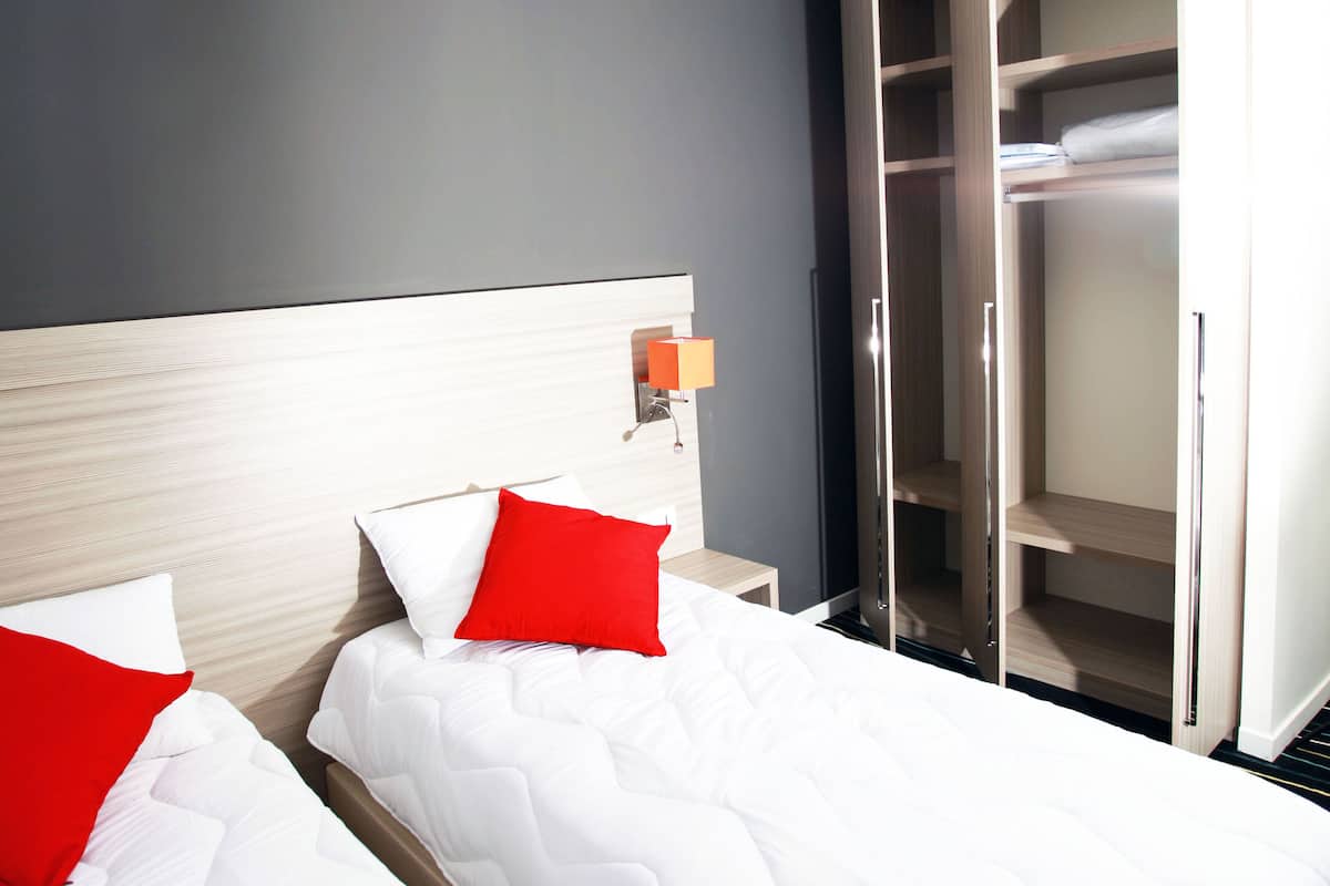 Une chambre d’hôtel avec deux lits, chacun agrémenté de draps blancs et d’oreillers rouges, une table de chevet avec une lampe et une armoire avec un miroir.