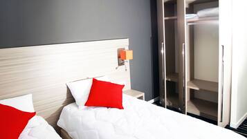 A hotel room with two beds, each with white bedding and red pillows, a bedside table with a lamp, and a wardrobe with a mirror.
